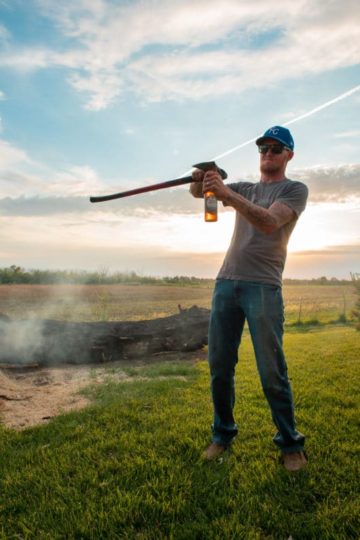 A man in a hat opens a beer with an axe