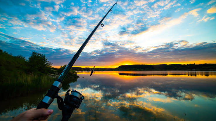 A person holding a fishing rod over a calm lake at sunrise or sunset, with colorful clouds reflecting in the water.